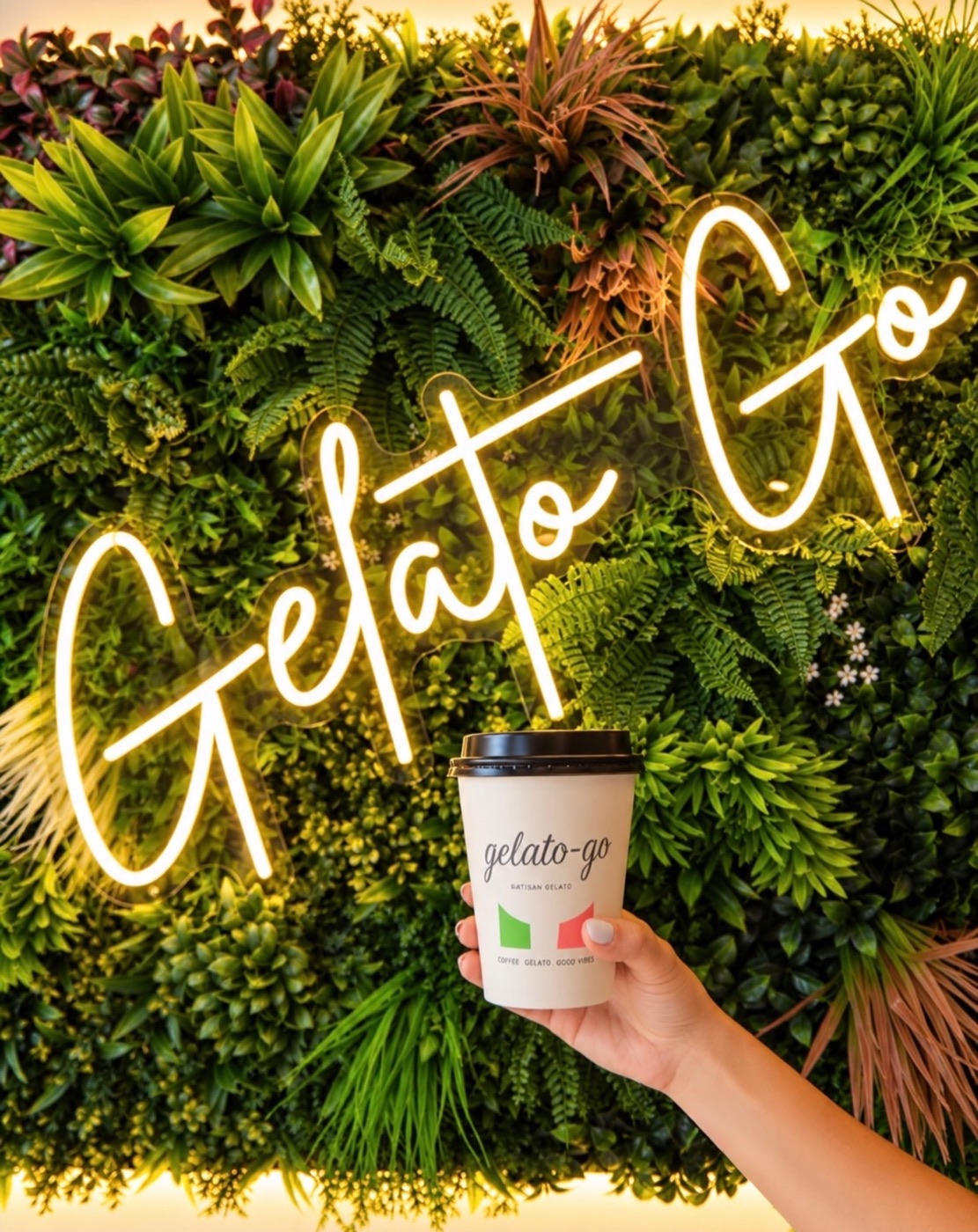 A Gelato-go cup held up in front of the neon Gelato Go sign on the green plant wall inside the Delray Beach shop